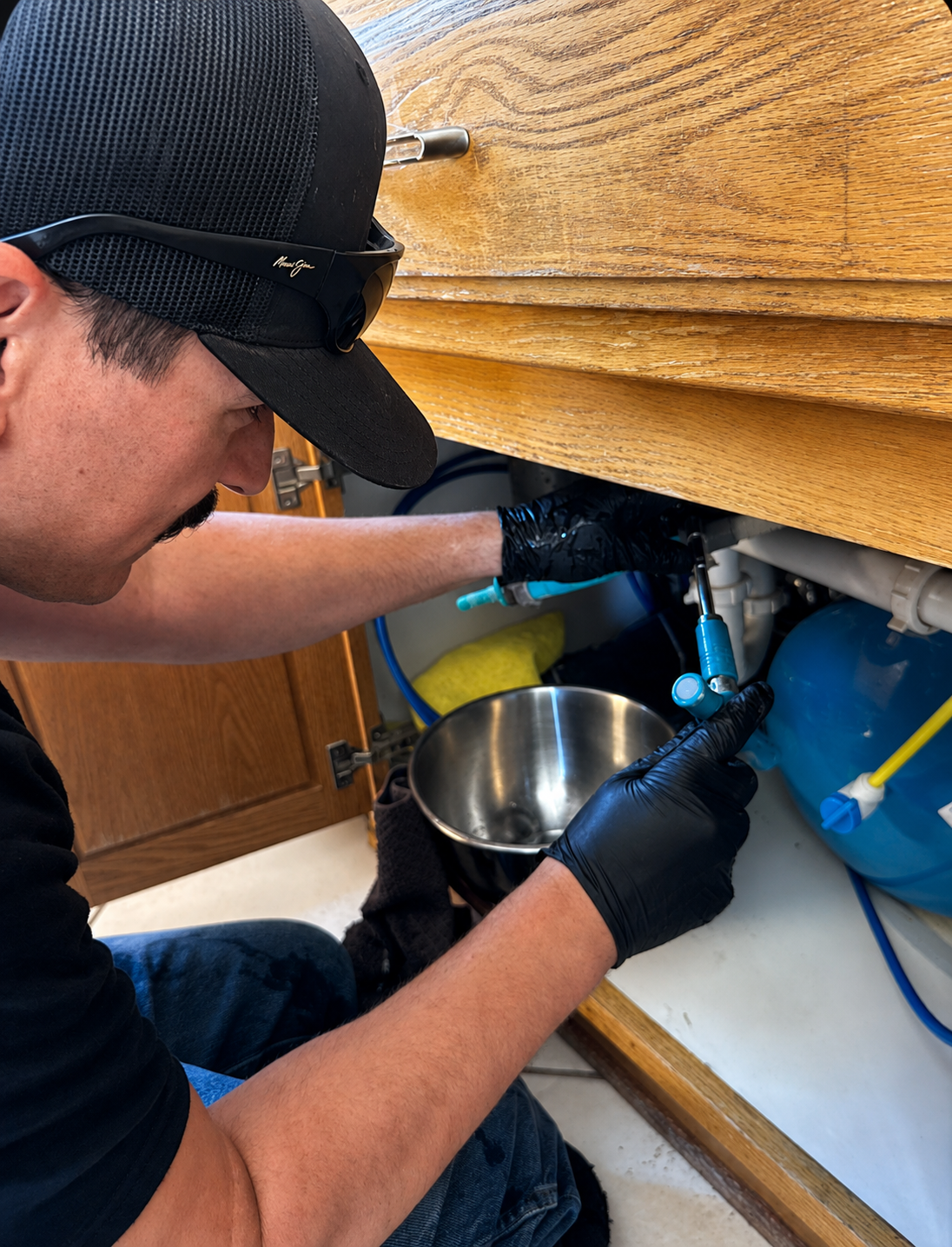 Plumber kneeling beside an open sink cabinet while servicing the supply connections underneath.
