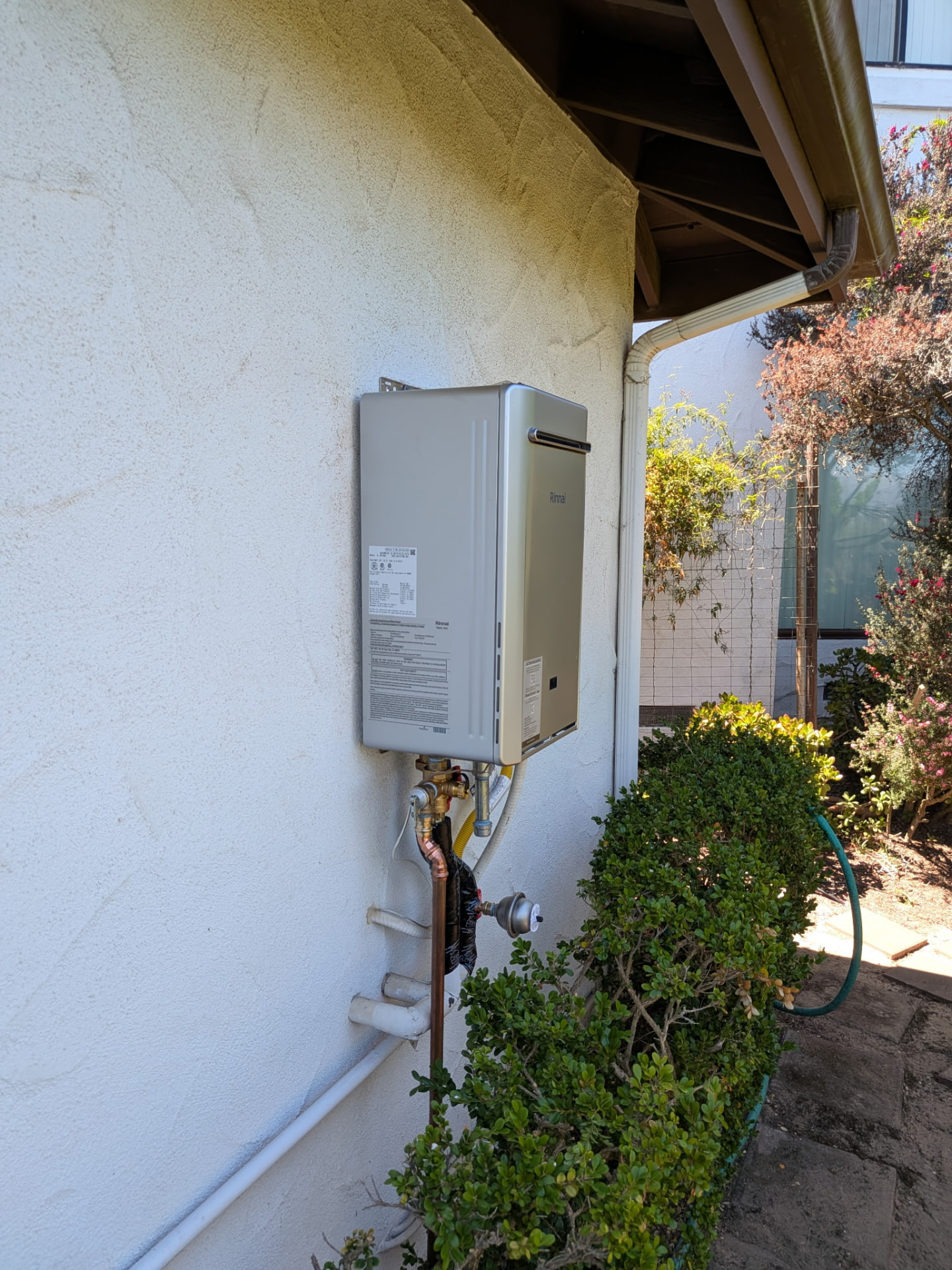Rinnai tankless water heater mounted on an exterior stucco wall under the eave with venting and gas connections.