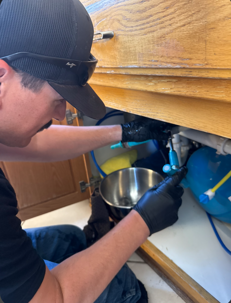 Plumber kneeling beside an open sink cabinet while servicing the supply connections underneath.
