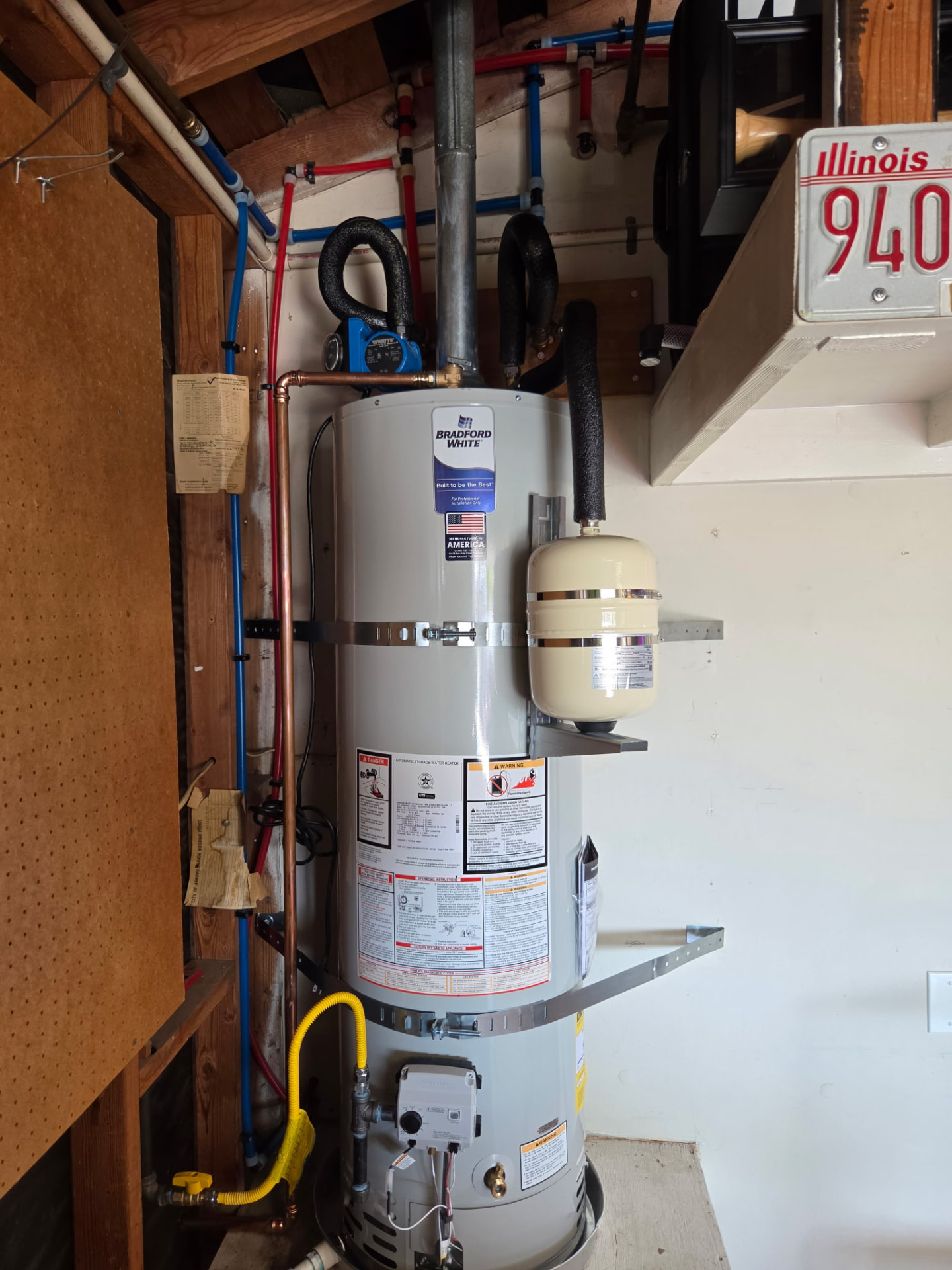 Bradford White gas water heater with expansion tank and safety pan installed in a garage utility space with copper, PEX and gas connections.
