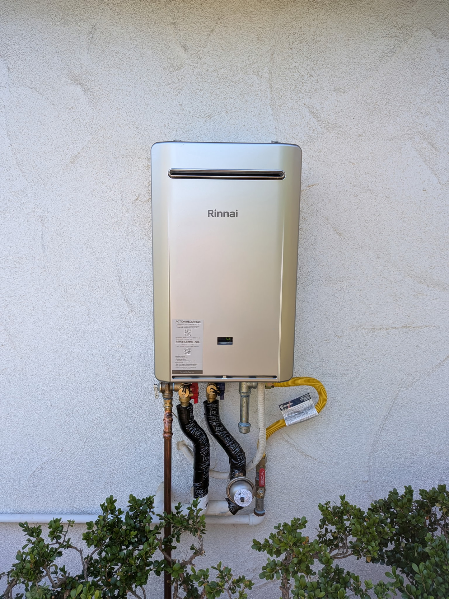 Front view of a wall-mounted Rinnai tankless water heater with isolation valves, gas connector and insulated water lines.